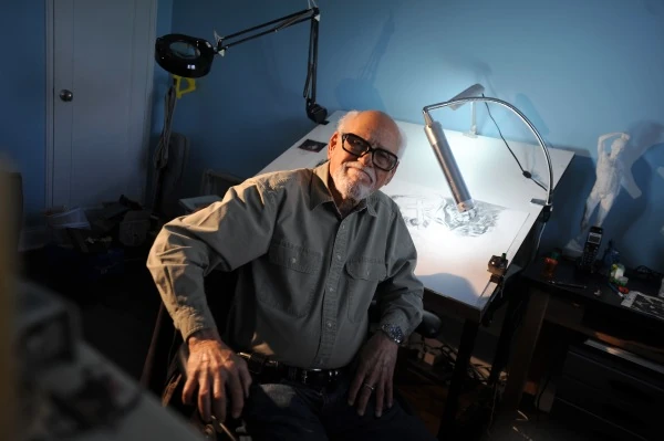 An elderly man sitting in a dimly lit workspace with drawing tools.